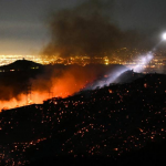 Incendio nocturno en la ladera de una colina con una ciudad iluminada al fondo.