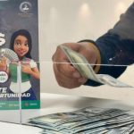 Persona depositando billetes en una caja transparente junto a un cartel promocional.