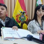 Dos personas sentadas en una mesa con banderas de fondo y micrófonos delante.