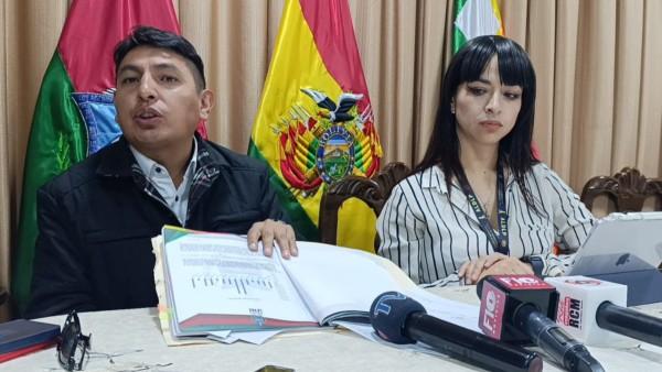 Dos personas sentadas en una mesa con banderas de fondo y micrófonos delante.