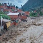 Río caudaloso pasando junto a casas de ladrillo, en un área montañosa.