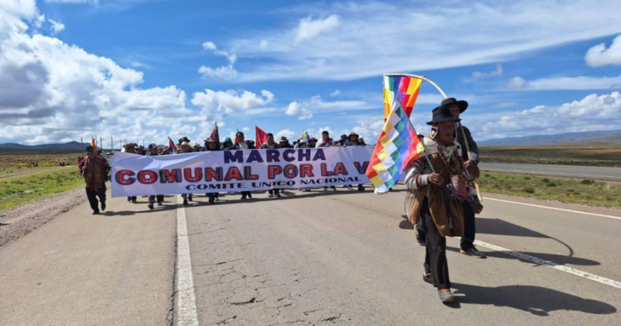 Un grupo de personas marchan por una carretera portando una gran pancarta y banderas coloridas.