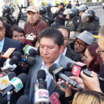 Grupo de personas rodeadas de micrófonos y reporteros con policías en el fondo.