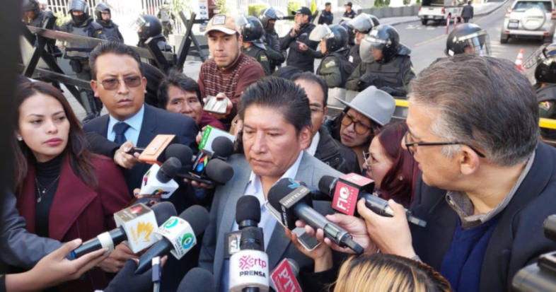 Grupo de personas rodeadas de micrófonos y reporteros con policías en el fondo.