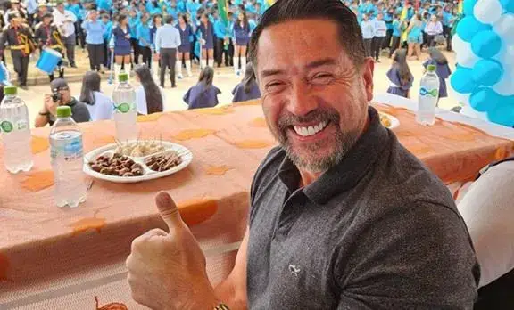 Un hombre sonriente con pulgar hacia arriba sentado en una mesa, con una multitud de personas al fondo.