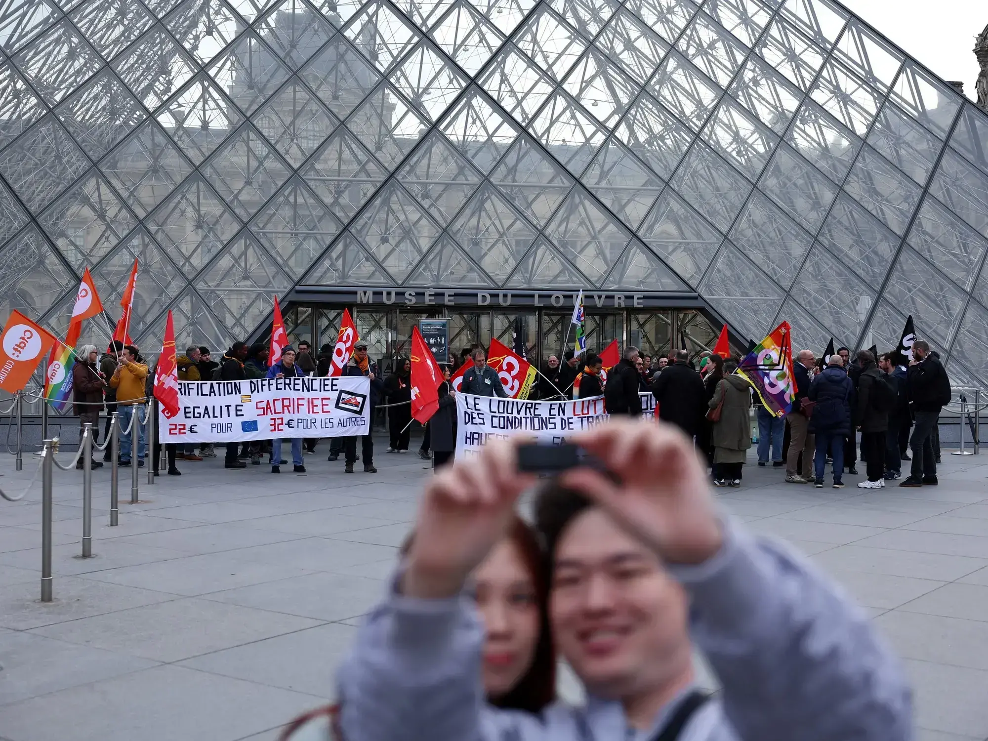 Dos turistas sacan una selfie en el Louvre, mientras los empleados del museo realizan una protesta.