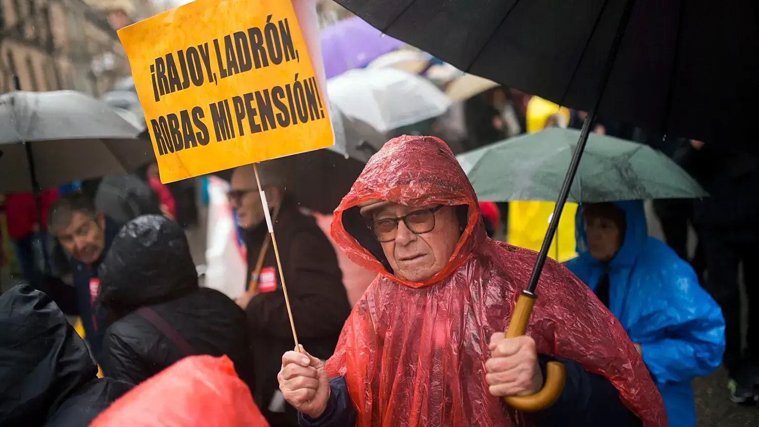 Un hombre con una pancarta que dice 'Rajoy ladrón, me robaste la pensión' camina bajo la lluvia durante una protesta de pensi