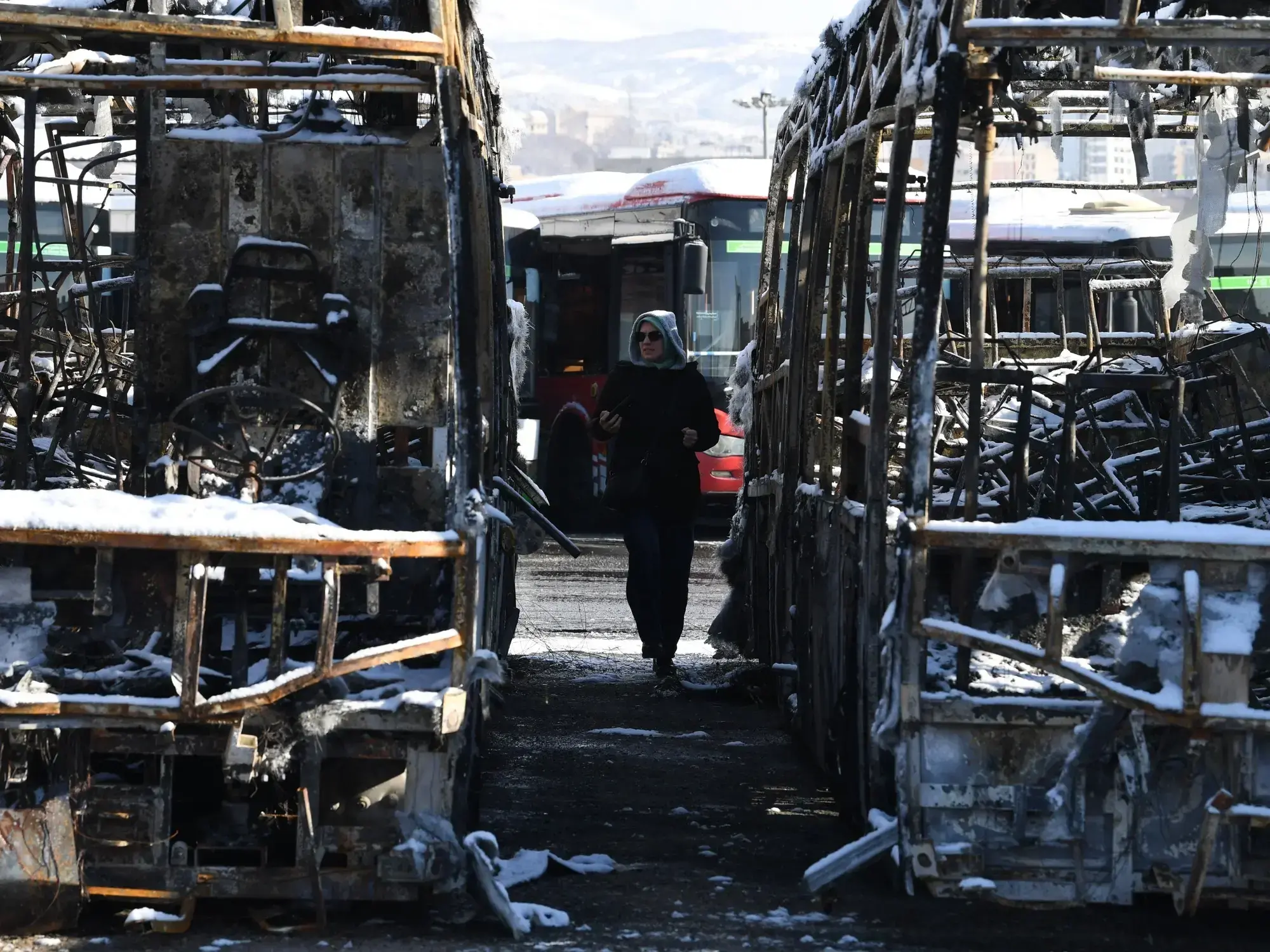 Una mujer camina entre dos colectivos destruidos tras las protestas en Irán.