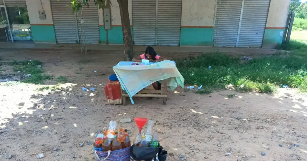 Niña haciendo tarea escolar en una mesa junto a recipientes con gasolina.