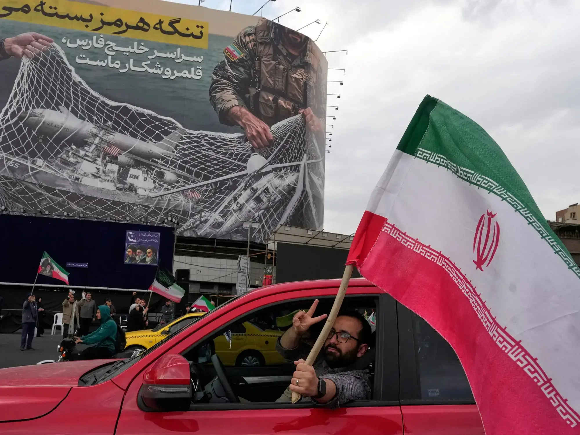 Un hombre celebra con una bandera de Irán bajo un cartel que ridiculiza a las fuerzas de Estados Unidos en Teherán.
