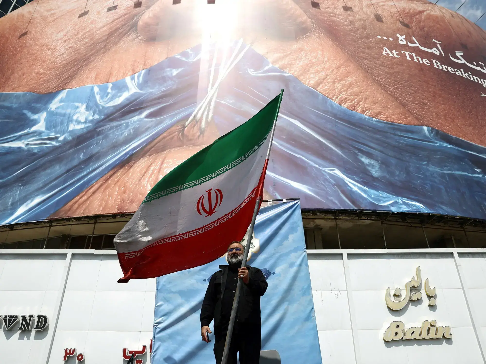 Un hombre sostiene la bandera de Irán frente a un cartel que muestra a Donald Trump amordazado.