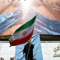 Un hombre sostiene la bandera de Irán frente a un cartel que muestra a Donald Trump amordazado.